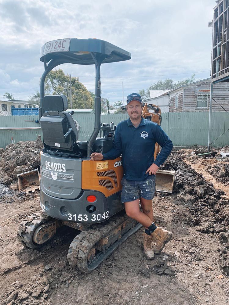 A Man is Standing Next to a Small Excavator on a Construction Site — Beards Roofing and Plumbing in Morayfield, QLD