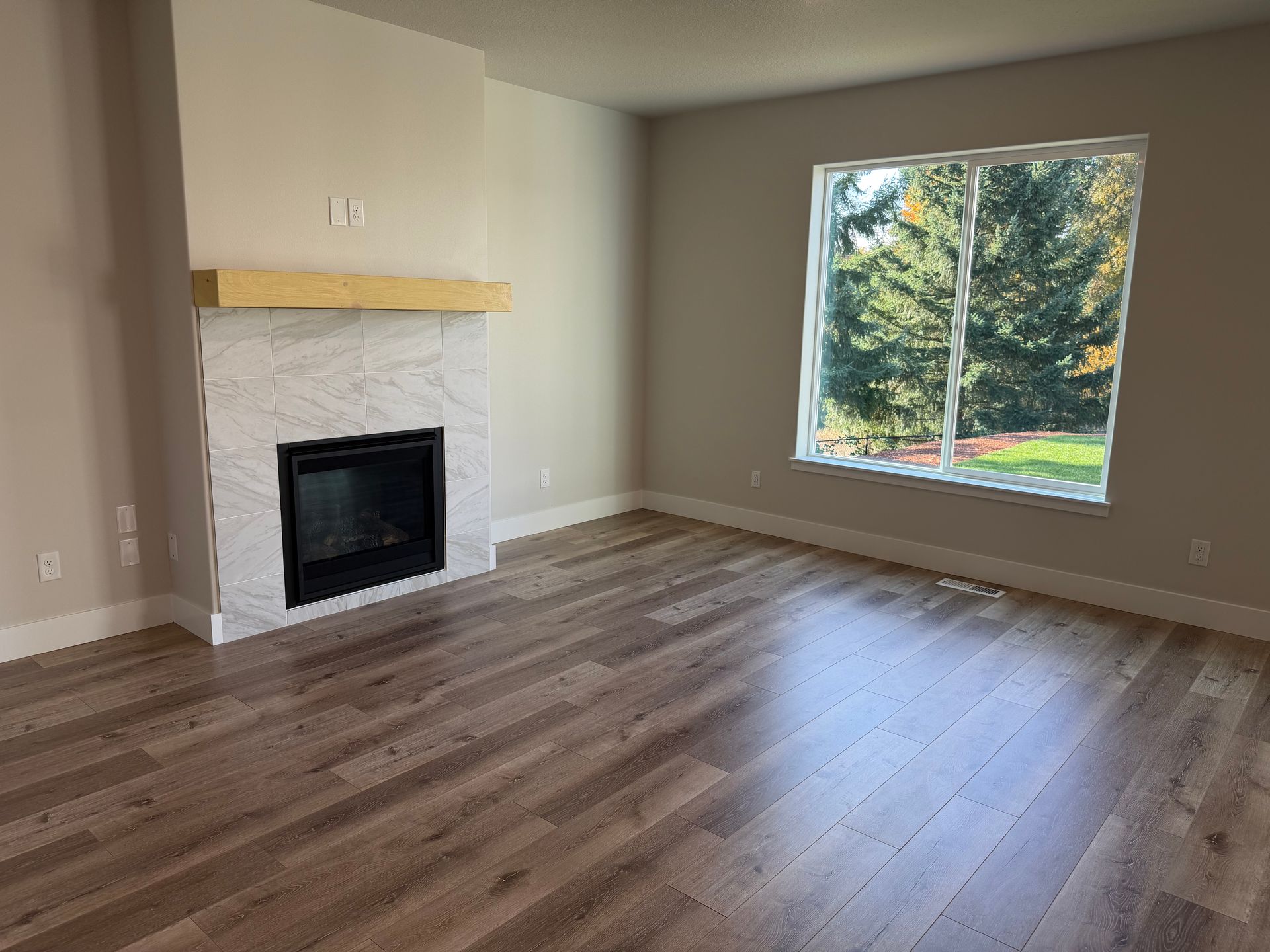 Empty living room with fireplace and large window overlooking trees, brown flooring.