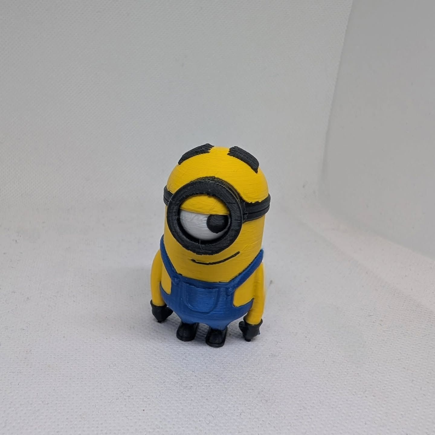 A small yellow Minion figurine in blue overalls standing on a white surface against a white backdrop.