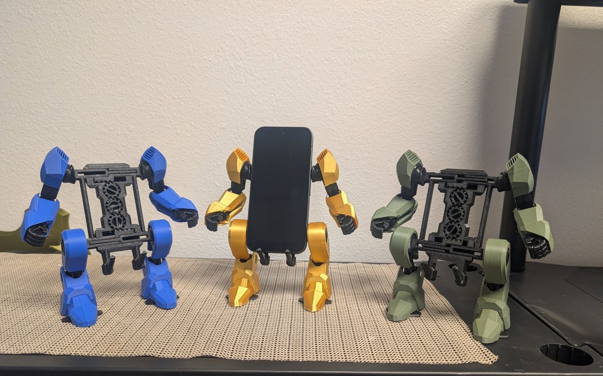 Three small robot-like figures in blue, yellow, and green standing on a table against a wall.