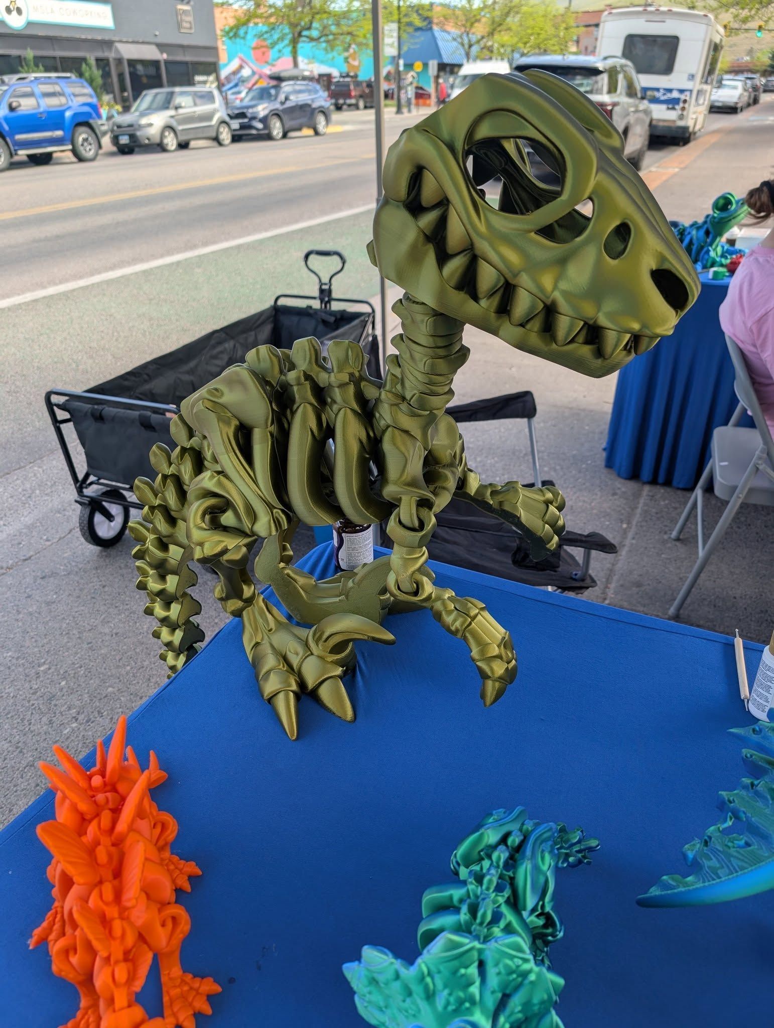 Green 3D-printed dinosaur skeleton figurine on a blue table at an outdoor market stall