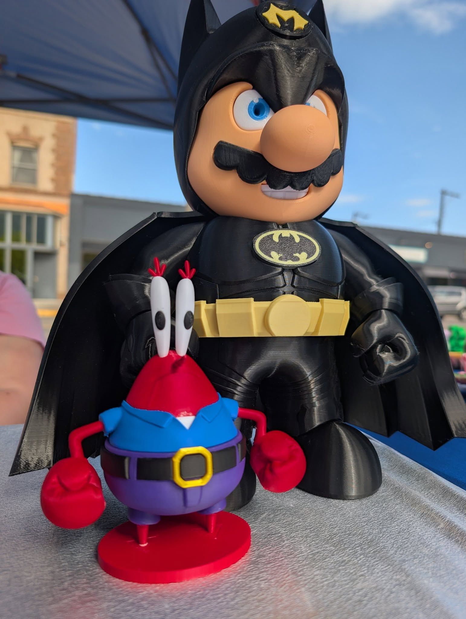 Batman figure standing behind a small blue-and-red toy on a table outdoors.