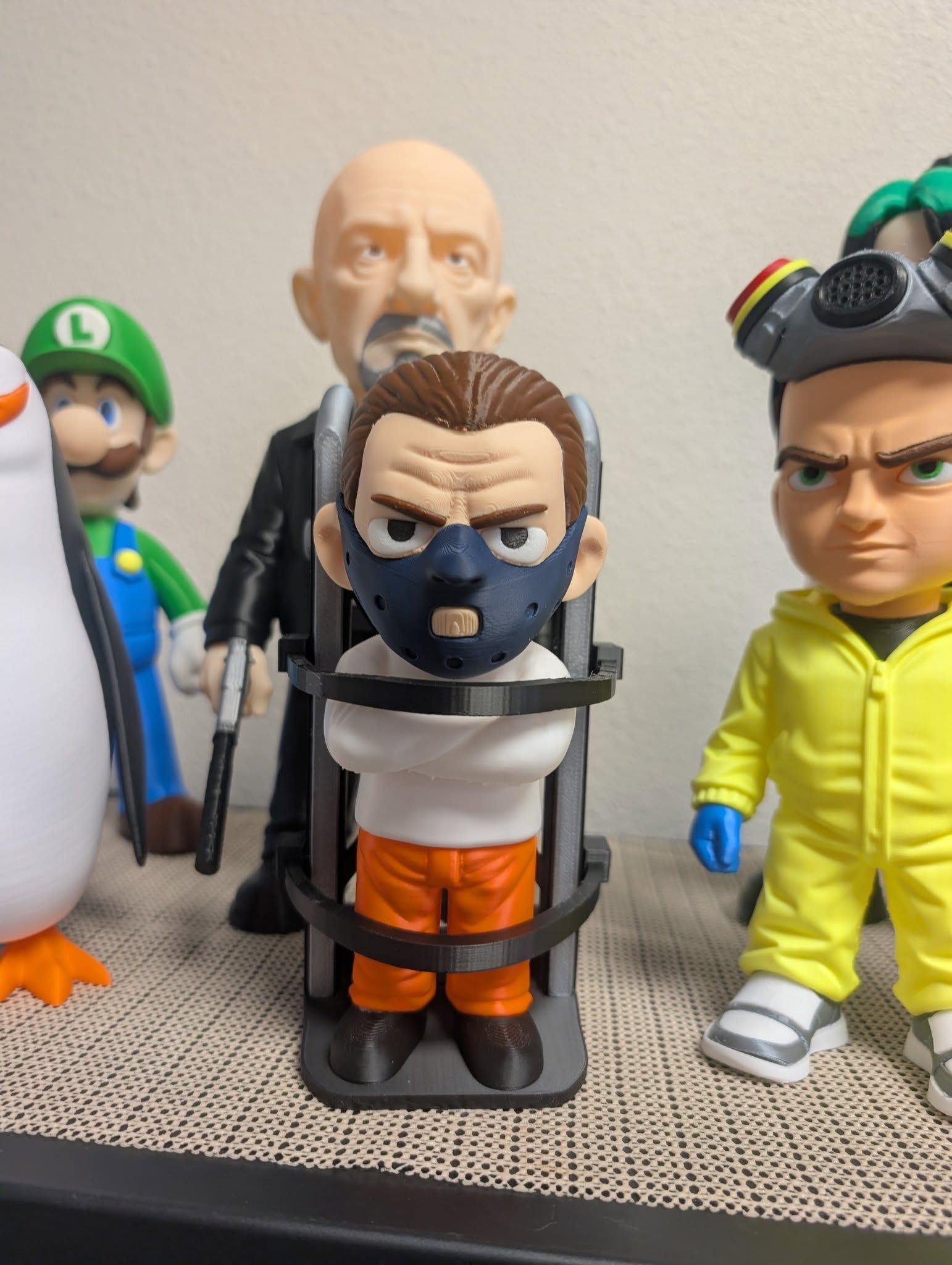 Toy figures lined up on a shelf, including Luigi, a masked figure in a cage, and a yellow-suited character.