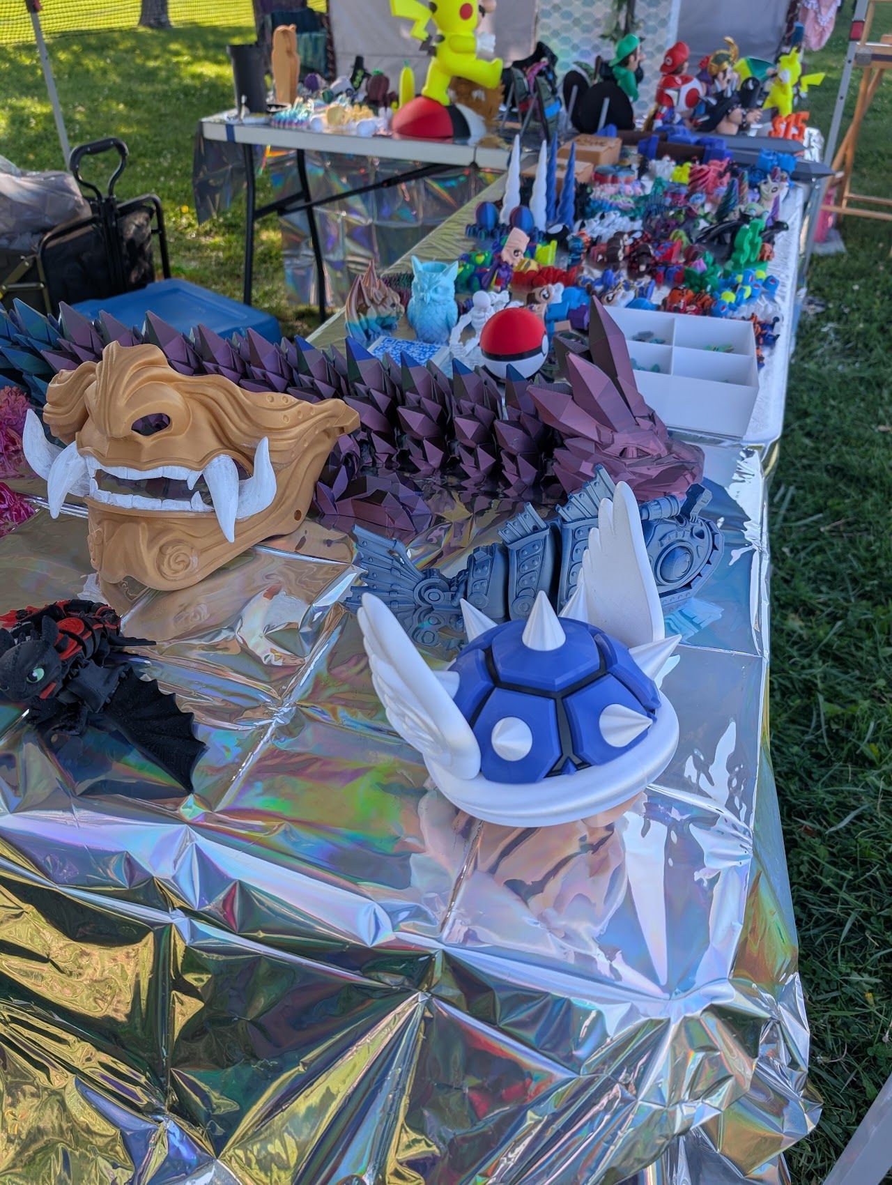 Table of colorful handmade craft masks and figurines on a shiny foil-covered display outdoors
