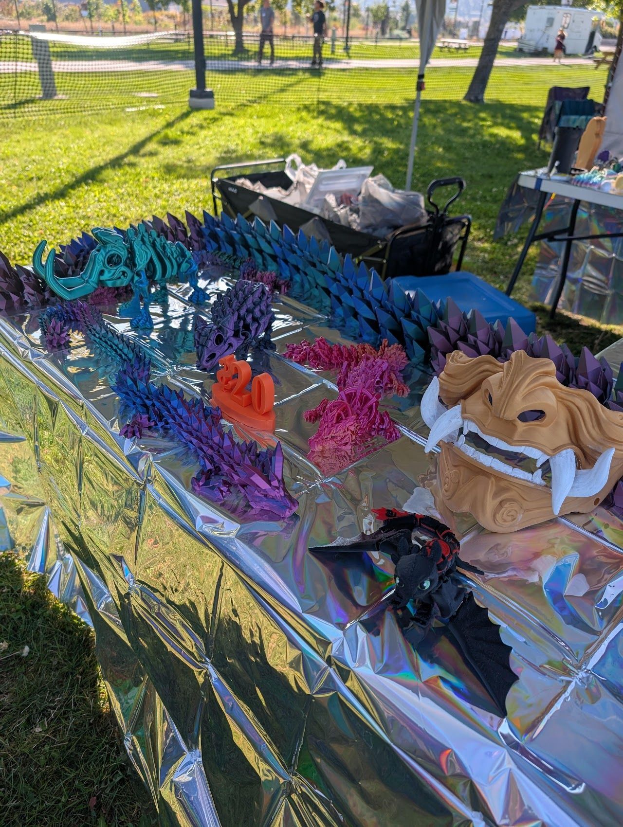 Colorful handmade crafts displayed on a foil-covered outdoor table in a sunny park.