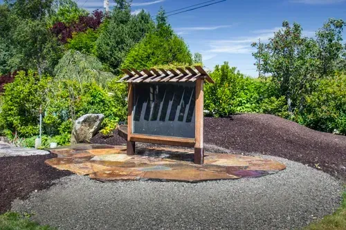 Outdoor kiosk sign with a shingled roof beside a gravel path and landscaped garden, under a blue sky