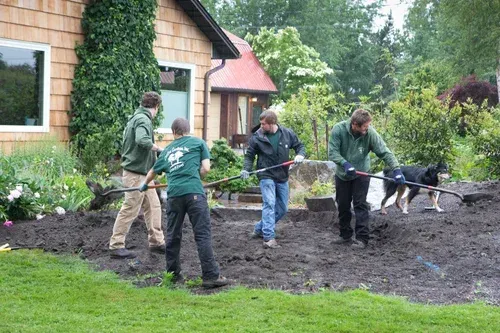 Four people shovel soil in a garden bed beside a house while a dog watches.