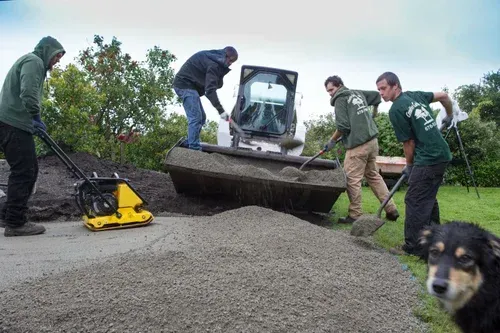 Workers with a skid steer spreading gravel on a road, with a dog in the foreground