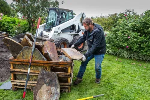Man in a black jacket loading rubble into a wooden skip beside a white skid steer in a grassy yard