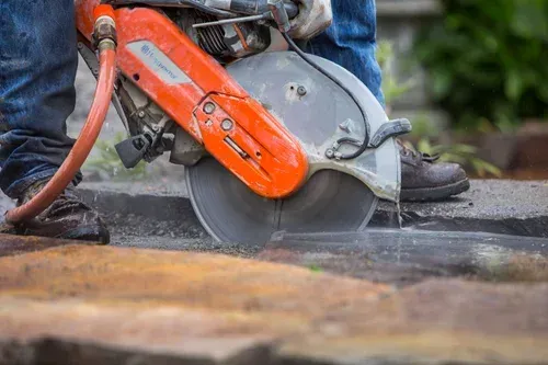 Worker using an orange circular saw to cut pavement, with dust and debris flying.