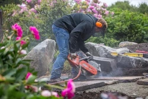 Worker using a chainsaw to cut stone outdoors, with dust and flowers nearby