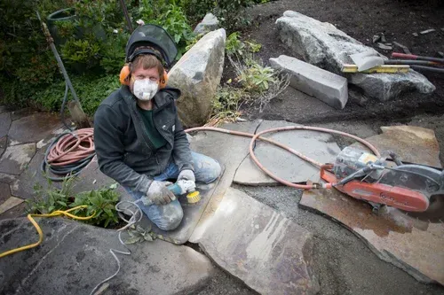 Worker kneeling in debris, wearing a mask and helmet, beside broken concrete and tools outdoors