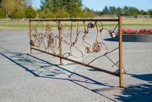 Rusty metal art fence casting shadows on a paved lot, with trees and flower beds in the background