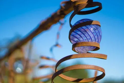 Blue glass ornament hanging from a spiral metal sculpture against a blurred sky background
