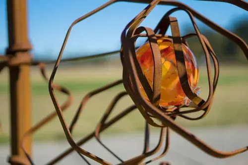 Close-up of a rusty wire sculpture with an amber glass center against a blurred outdoor sky and field.