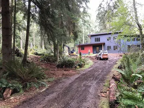 Forest driveway leading to a blue house with a red garage and parked vehicle