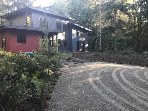 Curved gravel driveway beside a modern house with red and gray walls in a wooded setting