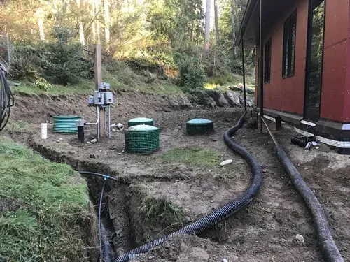 Outdoor utility area with green septic tanks, pipes, and a red building beside a wooded slope