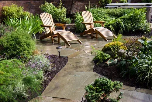 Two wooden lounge chairs on a stone patio in a lush green garden