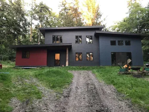 Modern dark-gray house with red side section, viewed from a gravel driveway and surrounded by trees