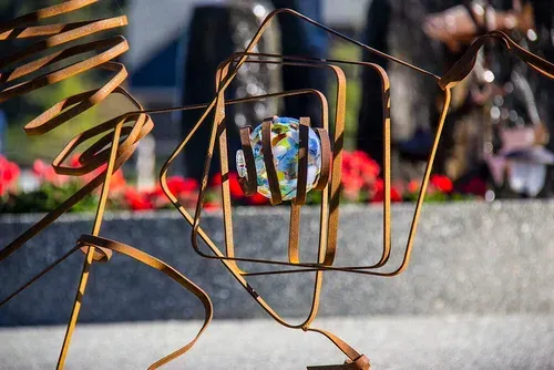 Abstract metal sculpture in front of blurred red flowers and a colorful glass sphere outdoors