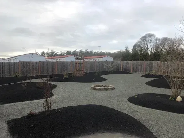 Backyard with gravel paths, mulched planting beds, young trees and a stone fire pit