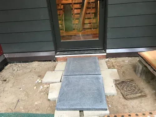 Temporary ramp leading to a doorway between dark siding, with dirt and concrete steps around it.