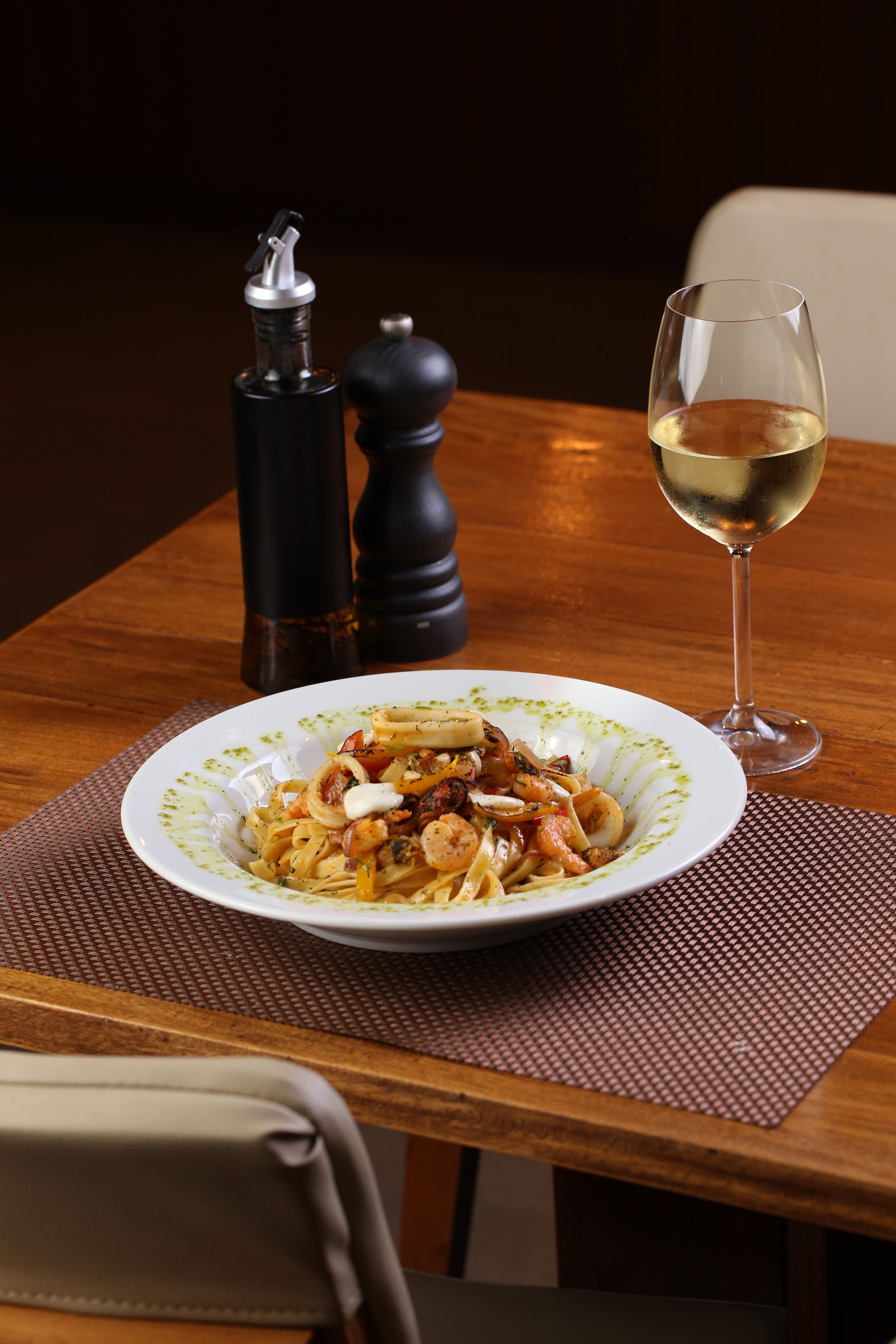Prato de massa com frutos do mar, taça de vinho, sal, pimenta e azeite sobre uma mesa de madeira.