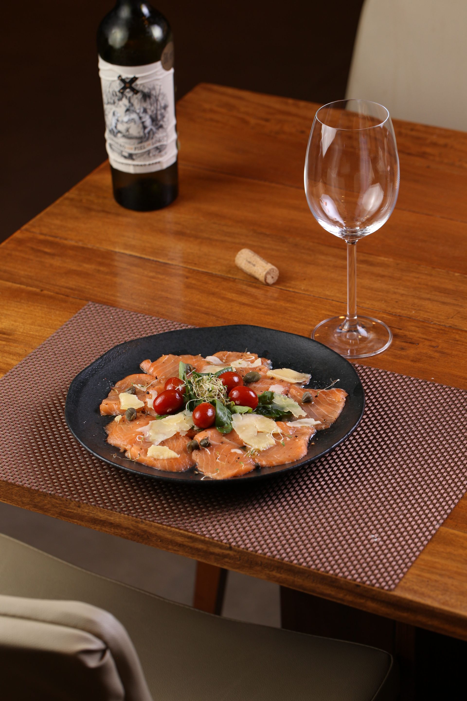 Prato de carpaccio com garrafa e taça de vinho sobre uma mesa de madeira.