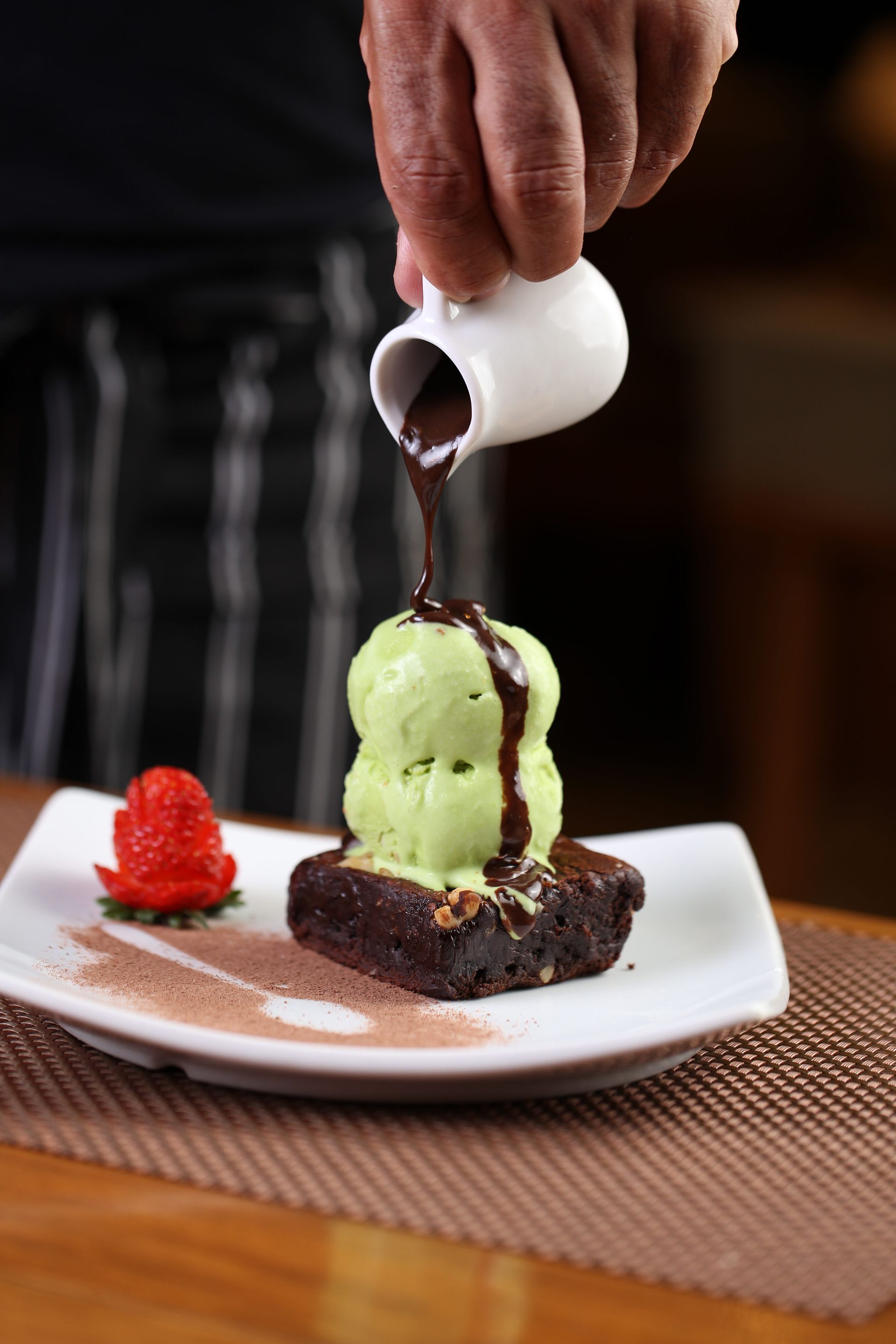 Chef despejando calda de chocolate sobre sorvete de matcha em um brownie com morango, em um prato branco.