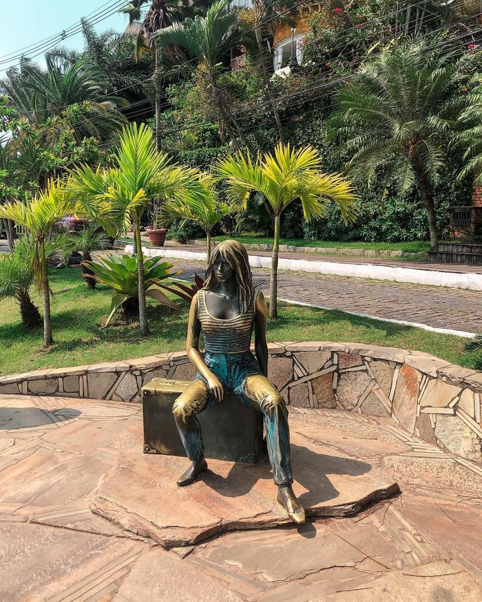 Estátua de uma pessoa sentada em um banco em um parque; tons de azul e dourado. Palmeiras e vegetação ao fundo.