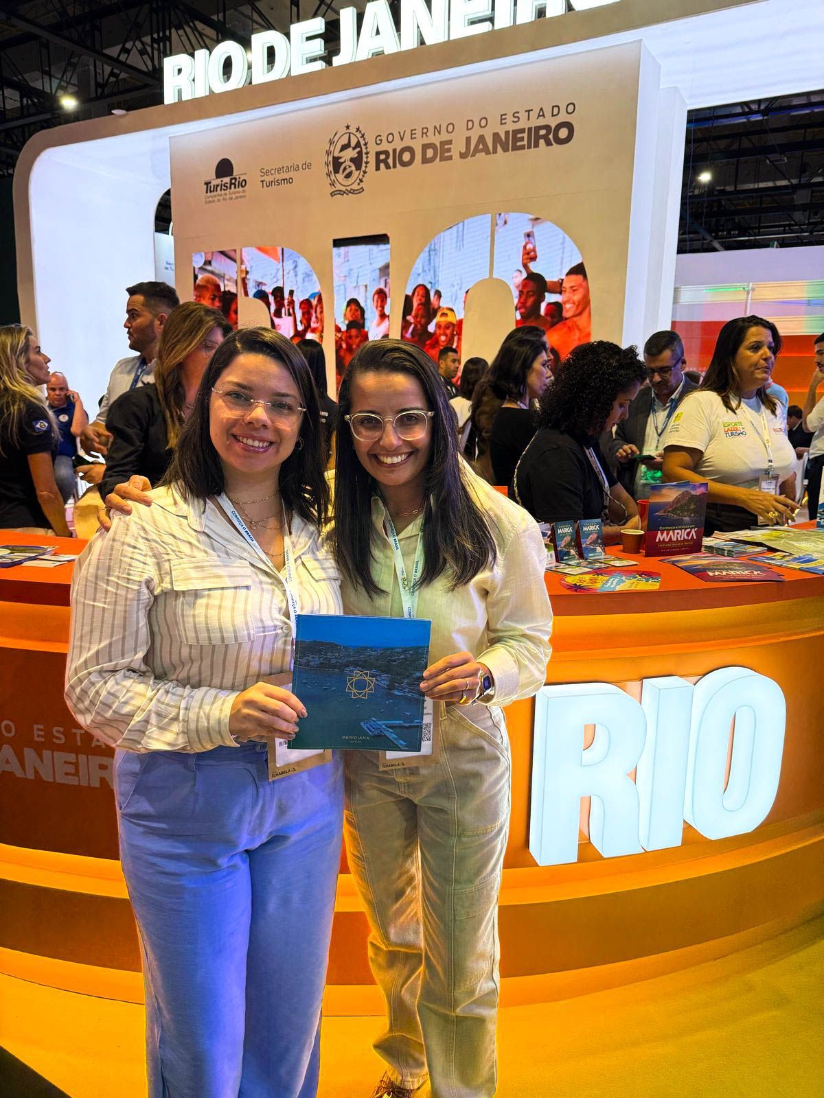 Duas mulheres sorridentes seguram um folheto em frente a um estande sobre o Rio de Janeiro em um evento turístico.