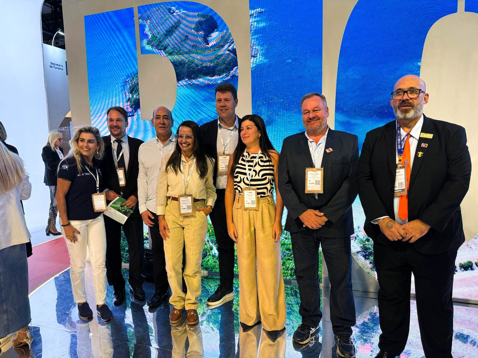 Grupo de pessoas em um estande de turismo do Rio de Janeiro com um fundo azul. As pessoas estão sorrindo e posando para fotos.