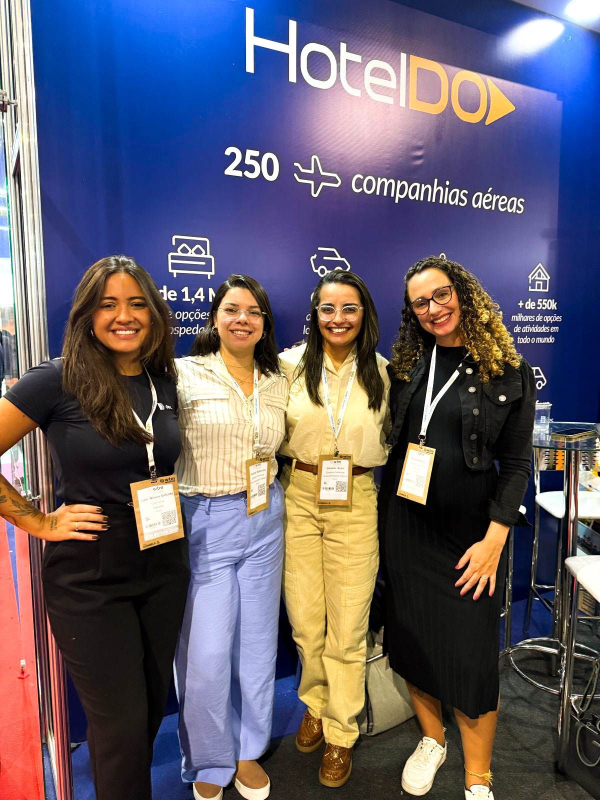 Quatro mulheres sorridentes posando em frente a uma faixa azul do HotelDO em um evento.