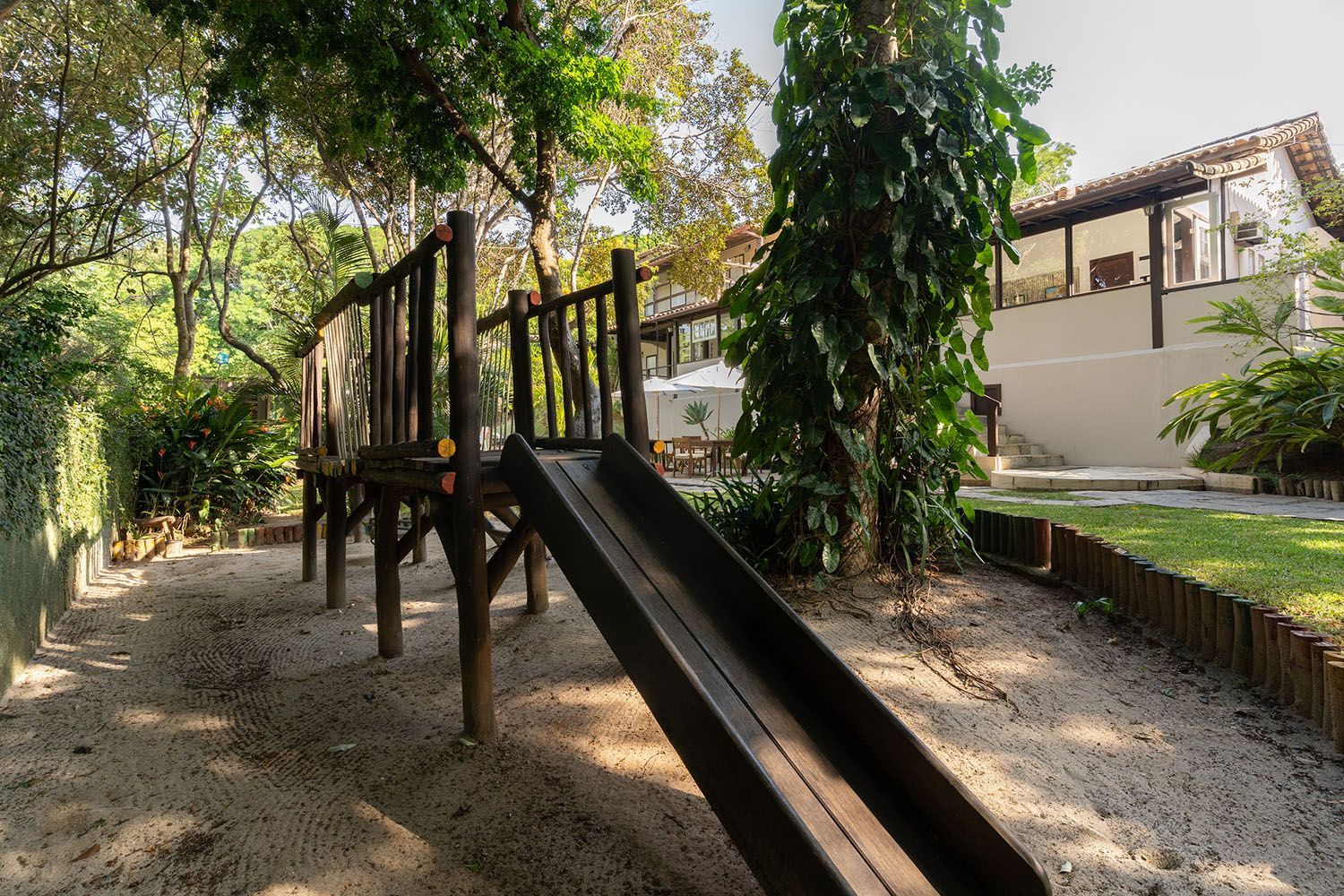 Tobogã de madeira em um parque infantil com areia, árvores e um prédio ao fundo.