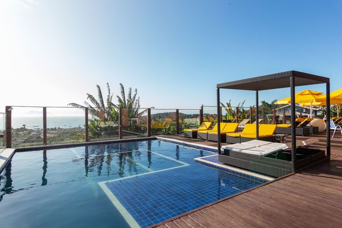 Piscina na cobertura com vista para o mar, espreguiçadeiras amarelas e uma cabana com sombra em um dia ensolarado.