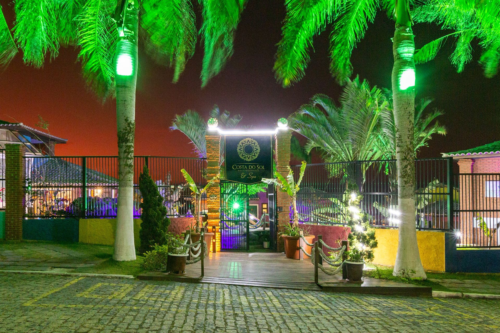 Entrada noturna para um local com palmeiras, luzes verdes e uma placa que diz 