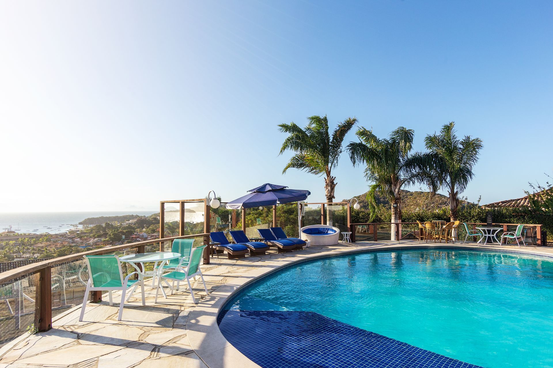 Piscina com espreguiçadeiras e palmeiras com vista para o oceano sob um céu límpido.