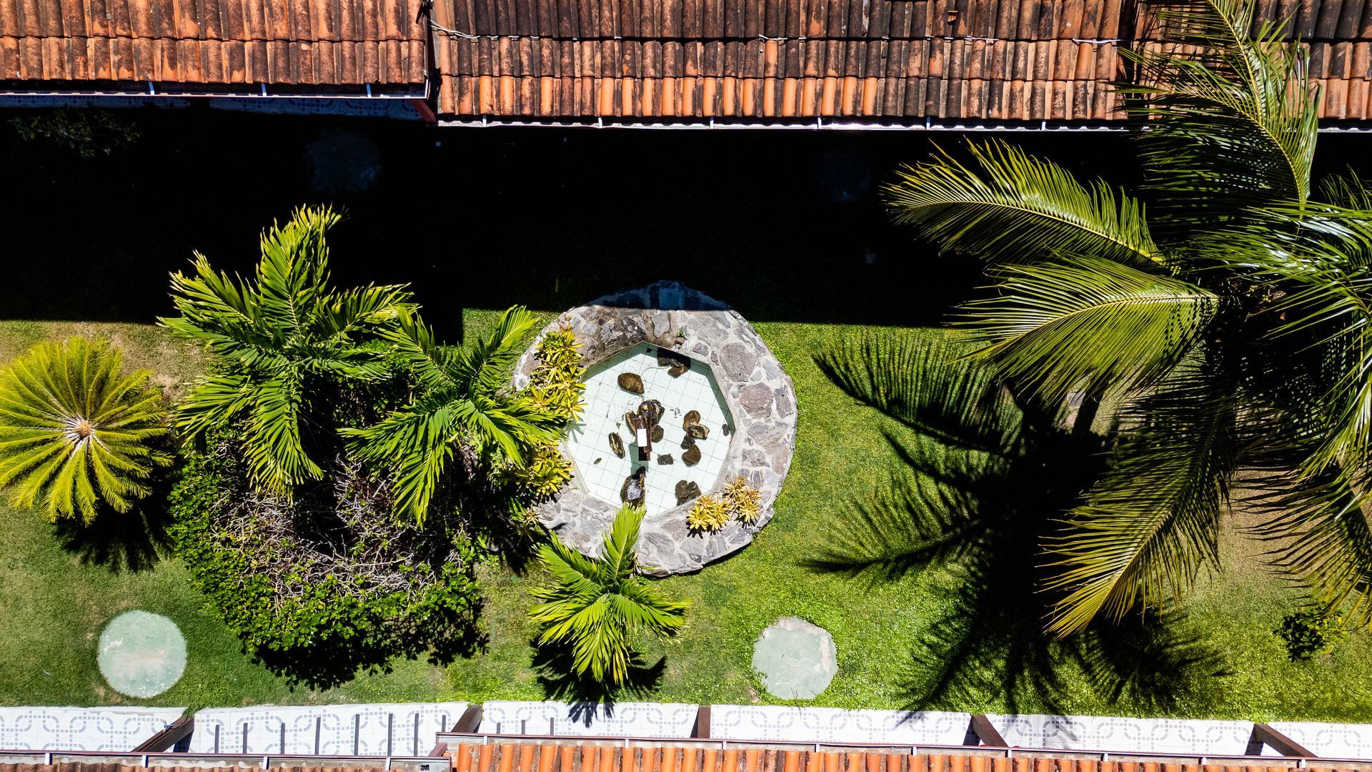 Vista aérea de um exuberante jardim verde com uma fonte de pedra e palmeiras, entre dois edifícios com telhados de telha vermelha.