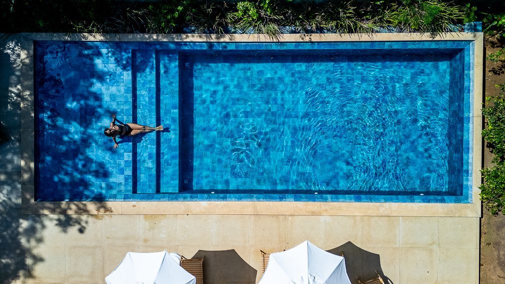 Uma pessoa flutua em uma piscina retangular azul brilhante, com degraus em uma das extremidades. Dois guarda-sóis brancos estão abaixo.