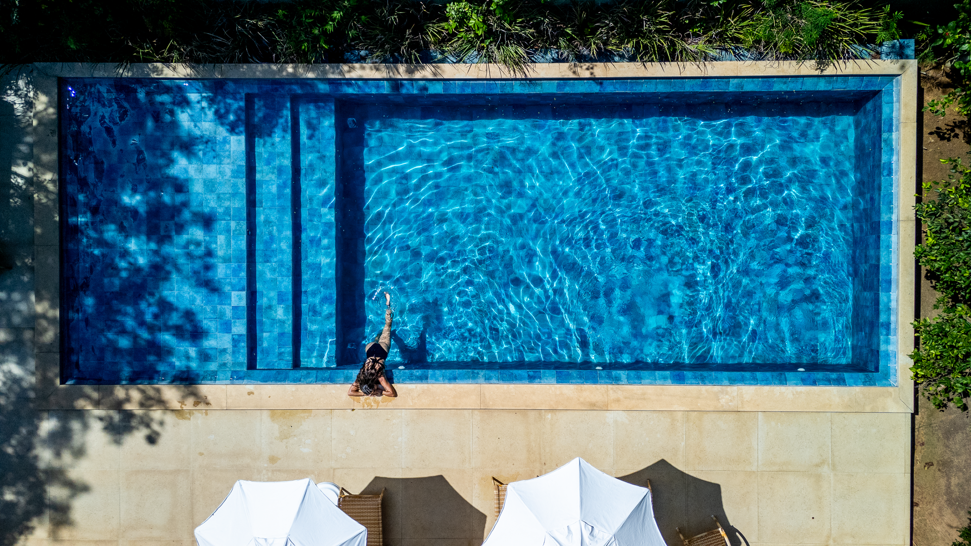 Pessoa em uma piscina. Vista aérea de uma piscina retangular azul com degraus e uma mulher nadando. Guarda-sóis brancos na lateral.