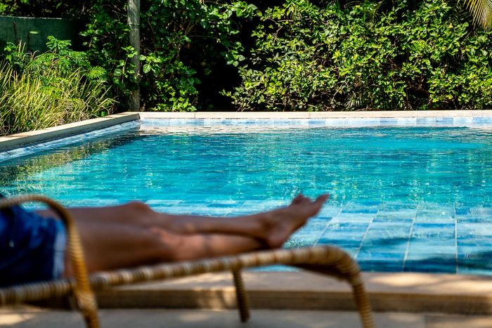 Pessoa relaxando em uma espreguiçadeira à beira de uma piscina com azulejos azuis, tendo como fundo uma exuberante folhagem verde.