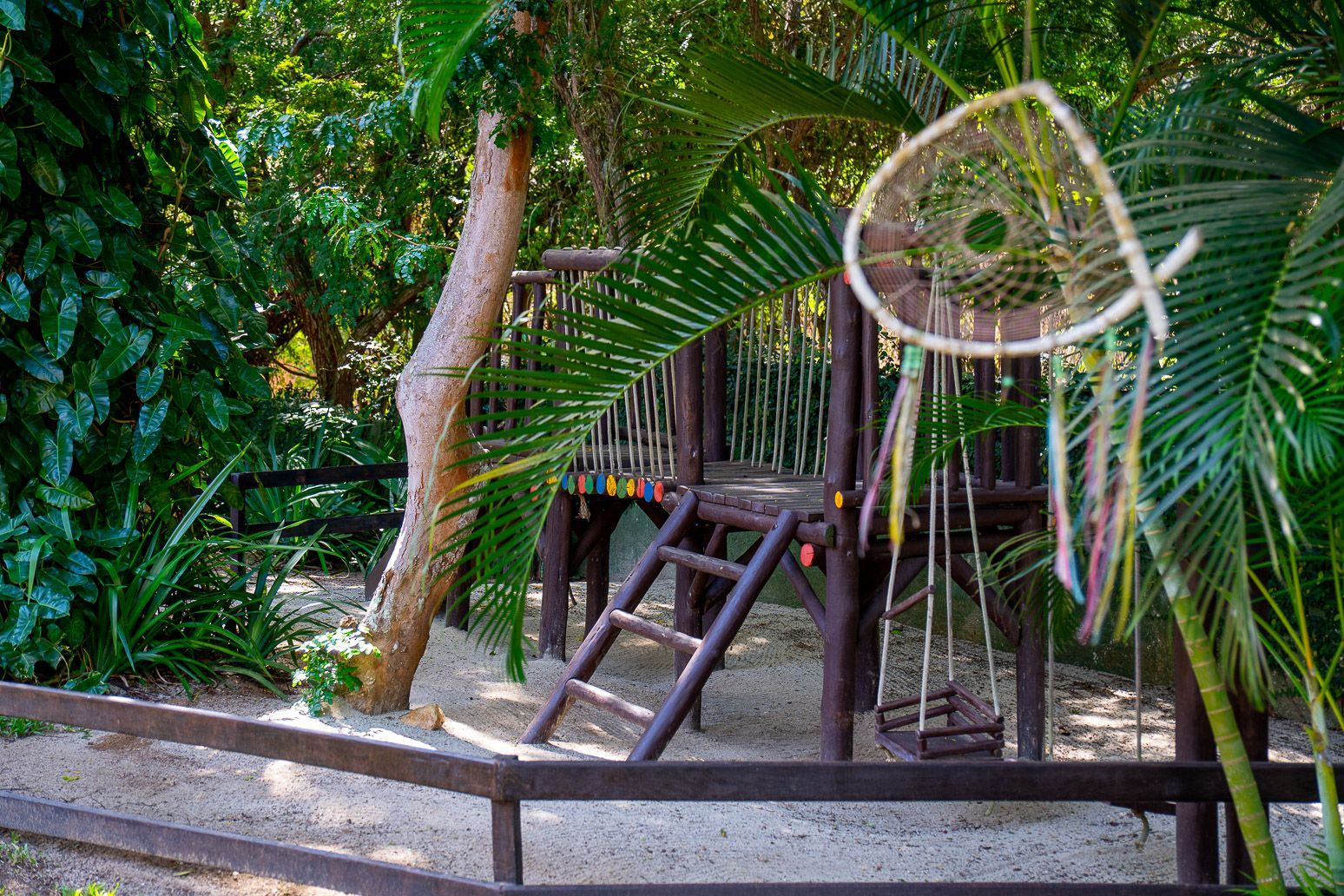 Parque infantil de madeira com escada e balanço, rodeado por folhagem verdejante e um apanhador de sonhos.
