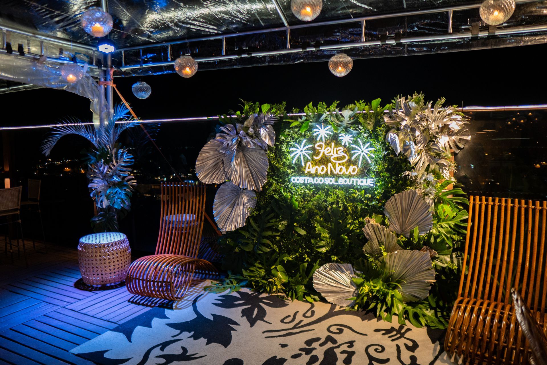 Evento festivo ao ar livre com cenário de folhagem verde, letreiro de néon, cadeiras de bambu e folhas prateadas decorativas.