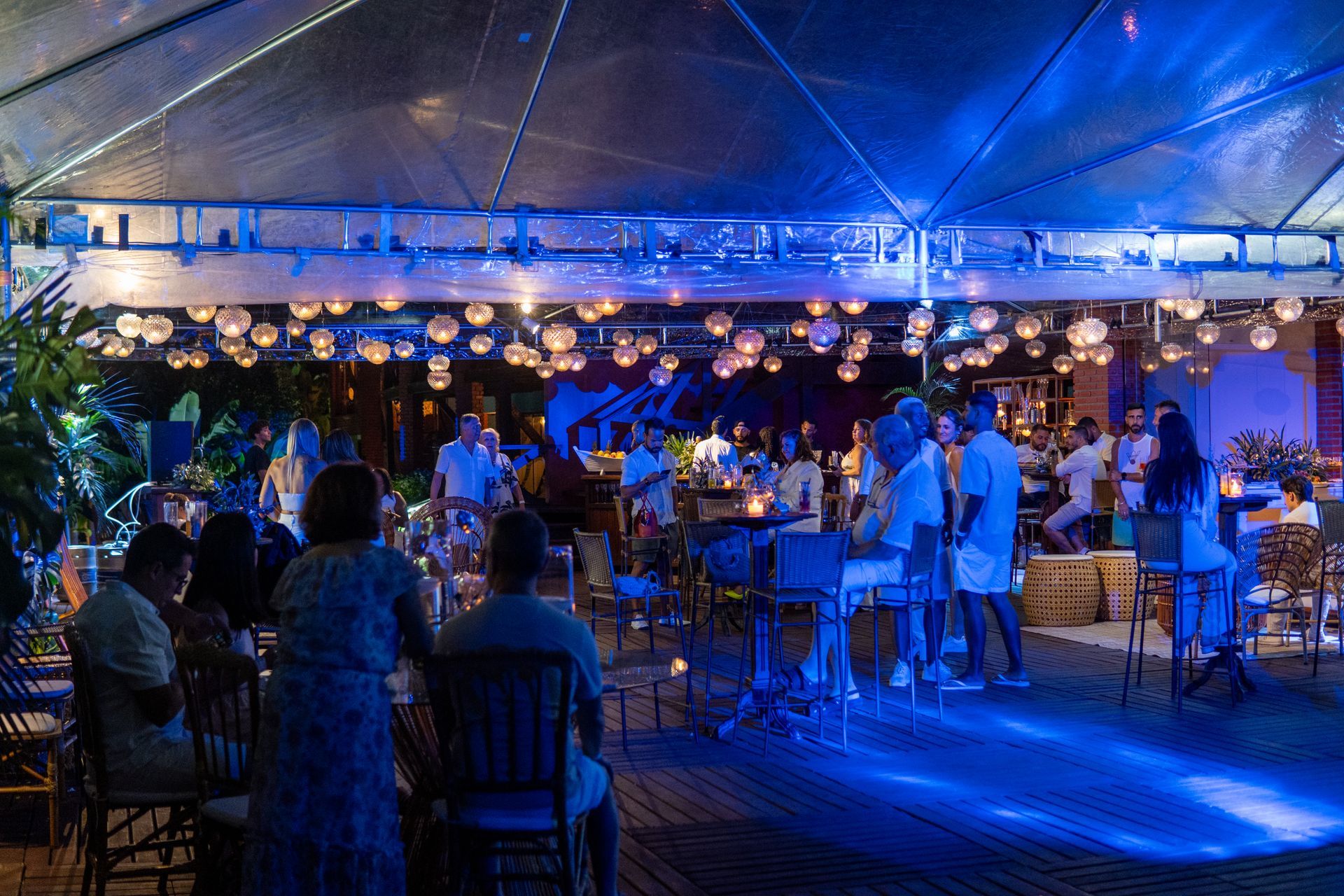 Pessoas em um evento ao ar livre sob um toldo iluminado, com iluminação azul e luzes esféricas.
