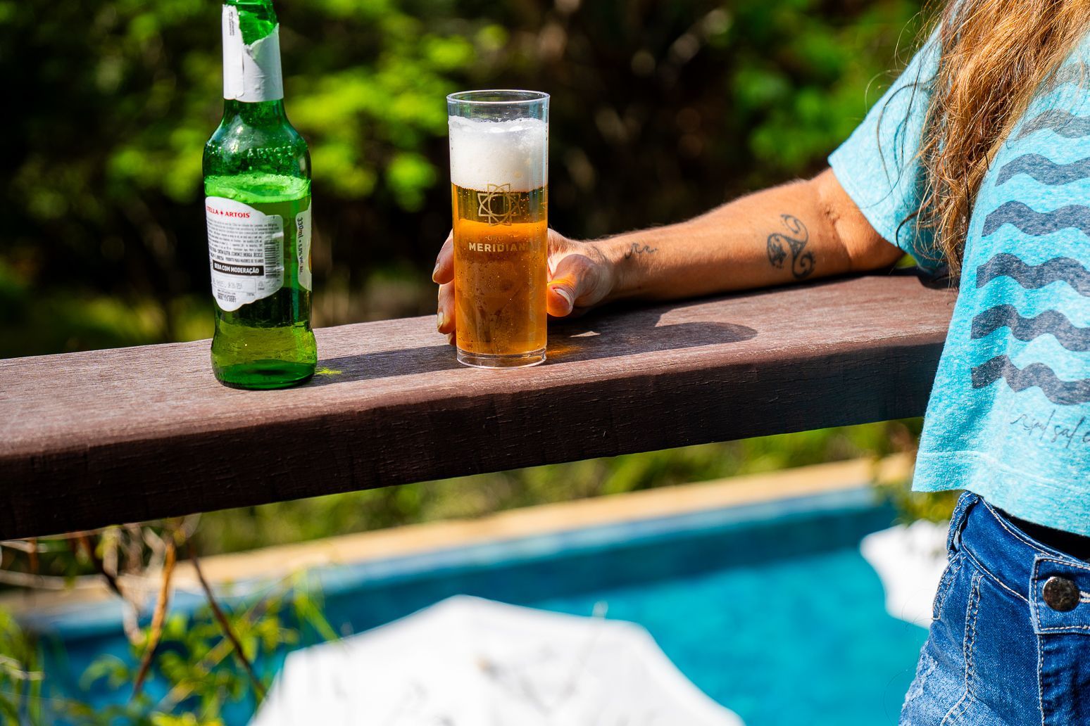 Garrafa de cerveja verde e copo alto de cerveja sobre um corrimão de madeira. Braço de pessoa segurando o copo, piscina ao fundo.