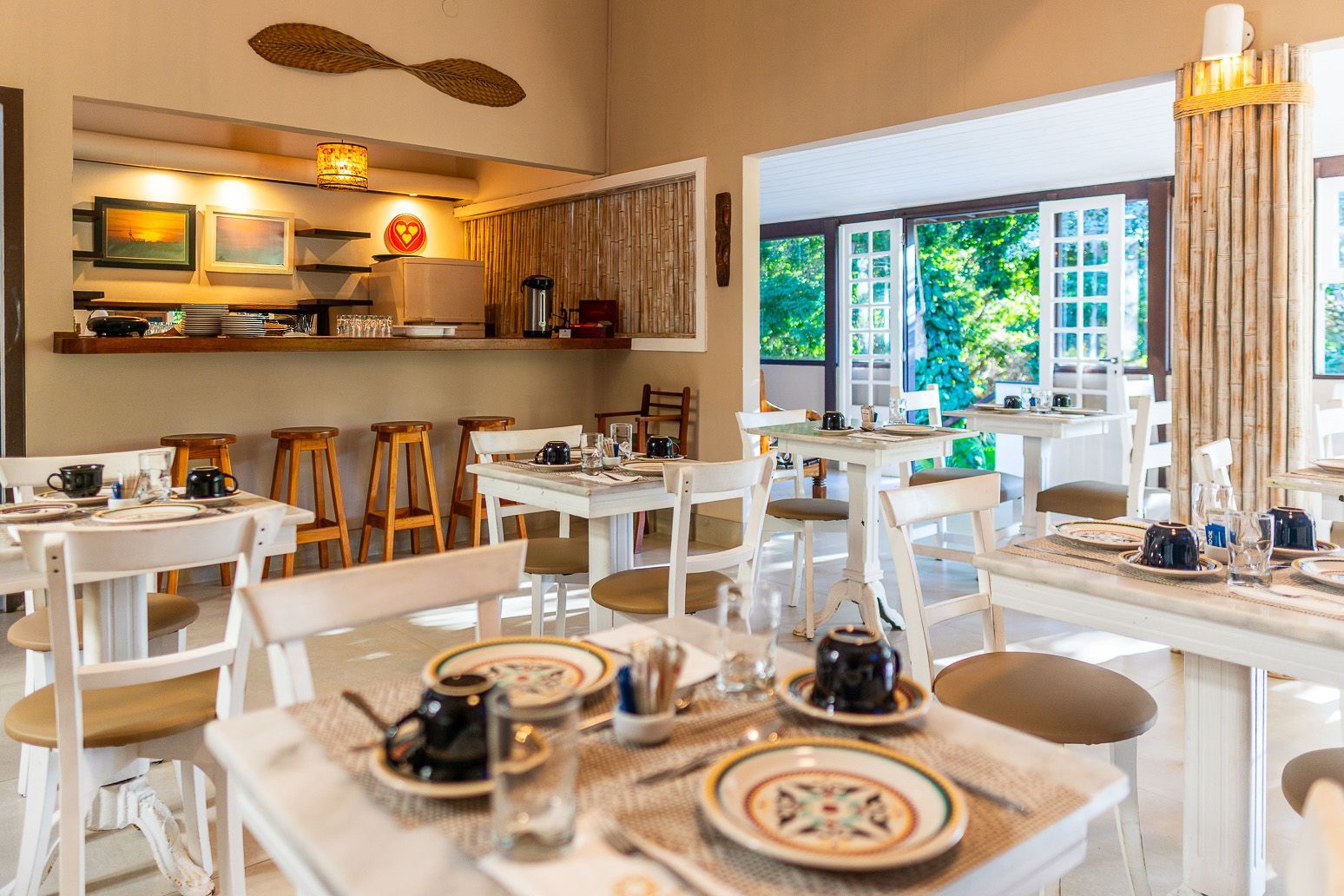 Sala de jantar iluminada com mesas, cadeiras e utensílios brancos. Um balcão de café da manhã está ao fundo.