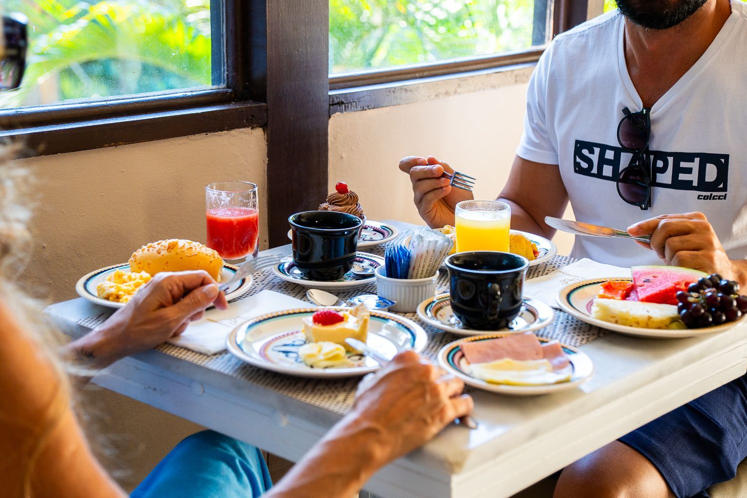Pessoas tomando café da manhã em uma mesa perto da janela, incluindo suco, frutas, ovos e café.