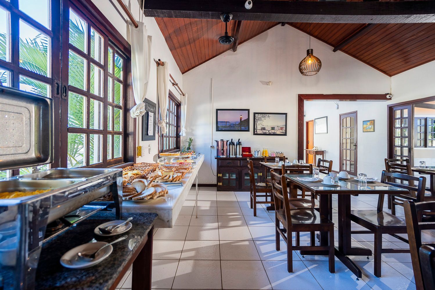 Buffet de café da manhã em uma sala de jantar com mesas de madeira, janelas e estações de serviço.