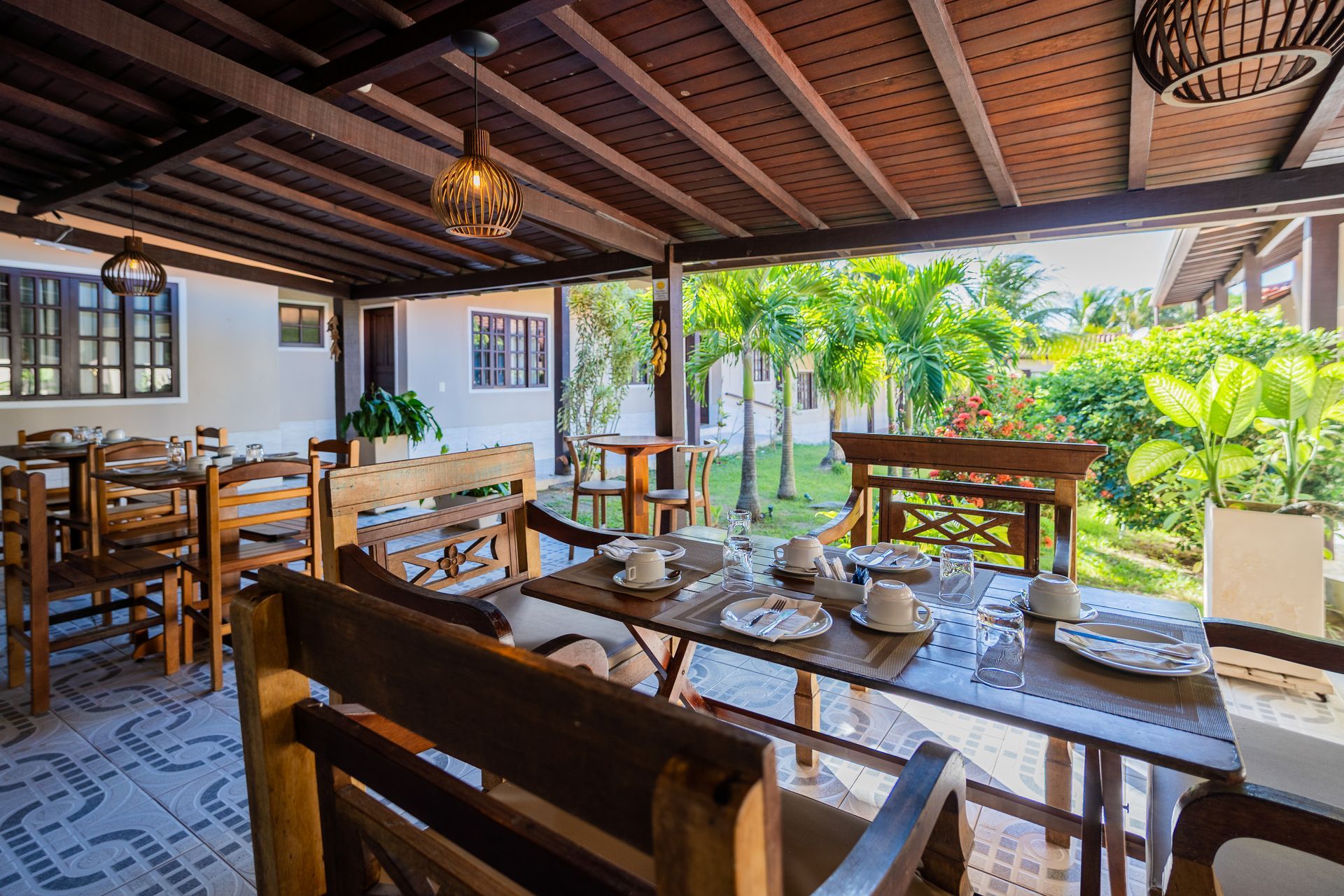 Pátio coberto com mesas postas para o café da manhã, com vista para um jardim tropical.